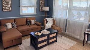 A living room with a brown sofa, a black coffee table, and a wicker chair. There are two windows with blinds, a floor lamp, and a view of the outside. - Inn on Turville Cottage, Sleeps 4, Waterfront (Berlin)