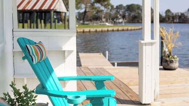A turquoise lounge chair with a striped pillow, a white porch, and a view of a lake with a dock and a building in the background. - Inn on Turville Cottage, Sleeps 4, Waterfront (Berlin)