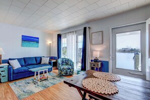 A living room with a blue sofa, a patterned armchair, a wooden table with coasters, and a view of a body of water through a large window.
