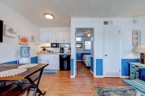 A compact kitchen with white cabinets, a microwave, and a water dispenser. A dining table with a placemat is in the foreground.