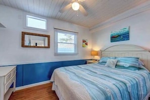 A bedroom with a bed, a nightstand, a ceiling fan, a window, and a painting on the wall.