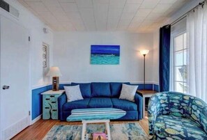 Ein Wohnzimmer mit einem blauen Sofa, einem gemusterten Sessel, einem gestreiften Couchtisch und einem Gemälde vom Meer.