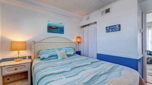 A bedroom with a bed, nightstand, lamp, and a sign indicating the direction to the beach.