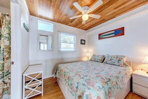 A bedroom with a wooden ceiling, a bed with a floral bedspread, a nightstand, a wall-mounted artwork, and a window with blinds.