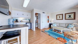 A spacious living area with a sofa, coffee table, and wicker chairs. A kitchen with white cabinets and a water dispenser is visible.