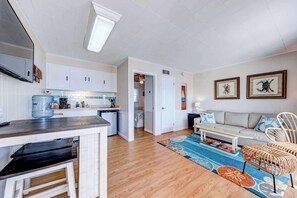 A spacious living area with a sofa, coffee table, and wicker chairs. A kitchen with white cabinets and a water dispenser is visible.