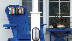 Une chaise Adirondack bleue avec un coussin à rayures, une colonne blanche ornée d’une grille métallique et une petite table sur laquelle est posée une plante en pot.