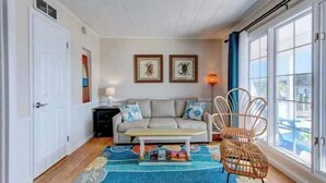 Un salon chaleureux avec un canapé, une table basse et un fauteuil en osier. Deux tableaux encadrés ornent le mur, et une fenêtre est agrémentée de rideaux.