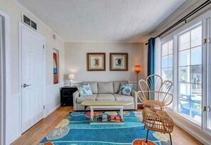 Un salon chaleureux avec un canapé, une table basse et un fauteuil en osier. Deux tableaux encadrés ornent le mur, et une fenêtre est agrémentée de rideaux.