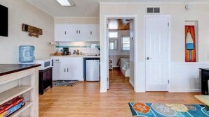 A compact kitchen with white cabinets, a microwave, and a water dispenser. A door leads to a bedroom with a bed and a window.