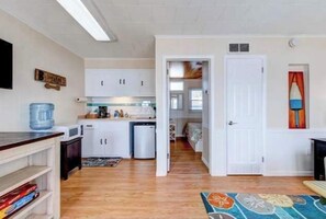 Cocina compacta con armarios blancos, un microondas y dispensador de agua. Una puerta conduce a un dormitorio con cama y ventana.