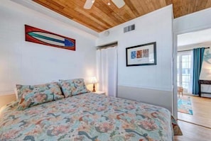 Un dormitorio con una colcha estampada de flores, un ventilador de techo, un cuadro enmarcado y una puerta que da a otra habitación.