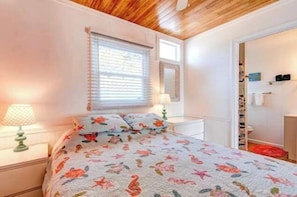 A bedroom with a bed, two bedside tables, a window with blinds, a mirror, and a door with a shoe rack.