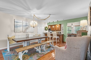 Uma sala de estar ampla com uma grande mesa de jantar de madeira, cadeiras brancas, um banco e um tapete colorido.