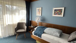 A hotel room with a bed, a chair, a bedside table, a fan, and two framed artworks on the wall. - Hotel Markgraf (Bad Bellingen)