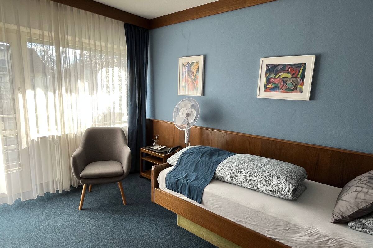 Une chambre d’hôtel avec un lit, une chaise, une table de chevet, un ventilateur et deux œuvres d’art encadrées accrochées au mur.