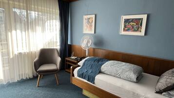 Hotelkamer met een bed, een stoel, een nachtkastje, een ventilator en twee ingekaderde kunstwerken aan de muur.