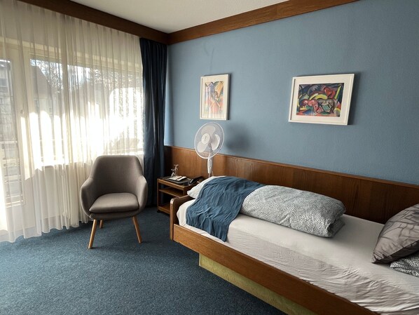 A hotel room with a bed, a chair, a bedside table, a fan, and two framed artworks on the wall. - Hotel Markgraf (Bad Bellingen)