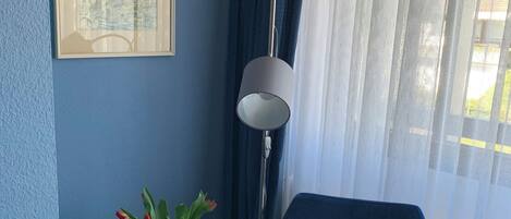 Egy szoba, melyben egy kék szék, egy kis asztal, rajta egy virágos vázában, egy lámpa és egy keretezett kép található a falon.