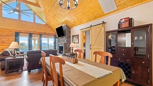 A spacious room with a high ceiling, wooden beams, a fireplace, a dining table with chairs, a TV, and a large window offering a view.
