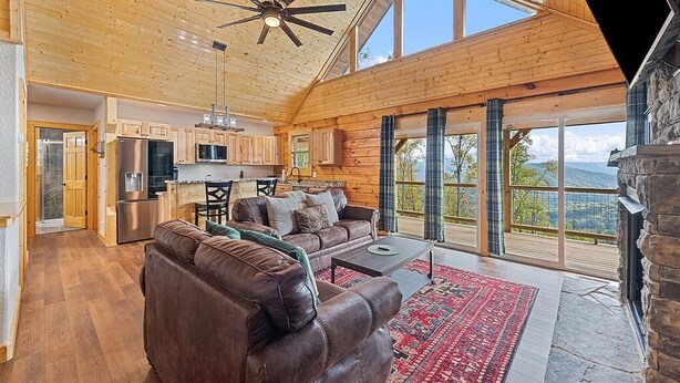 A spacious living area with a wooden ceiling, a stone fireplace, and large windows offering a view of the outdoors.