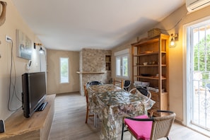 A dining area with a table covered in a floral tablecloth, chairs, a television on a wooden stand, a stone wall, and a bookshelf.