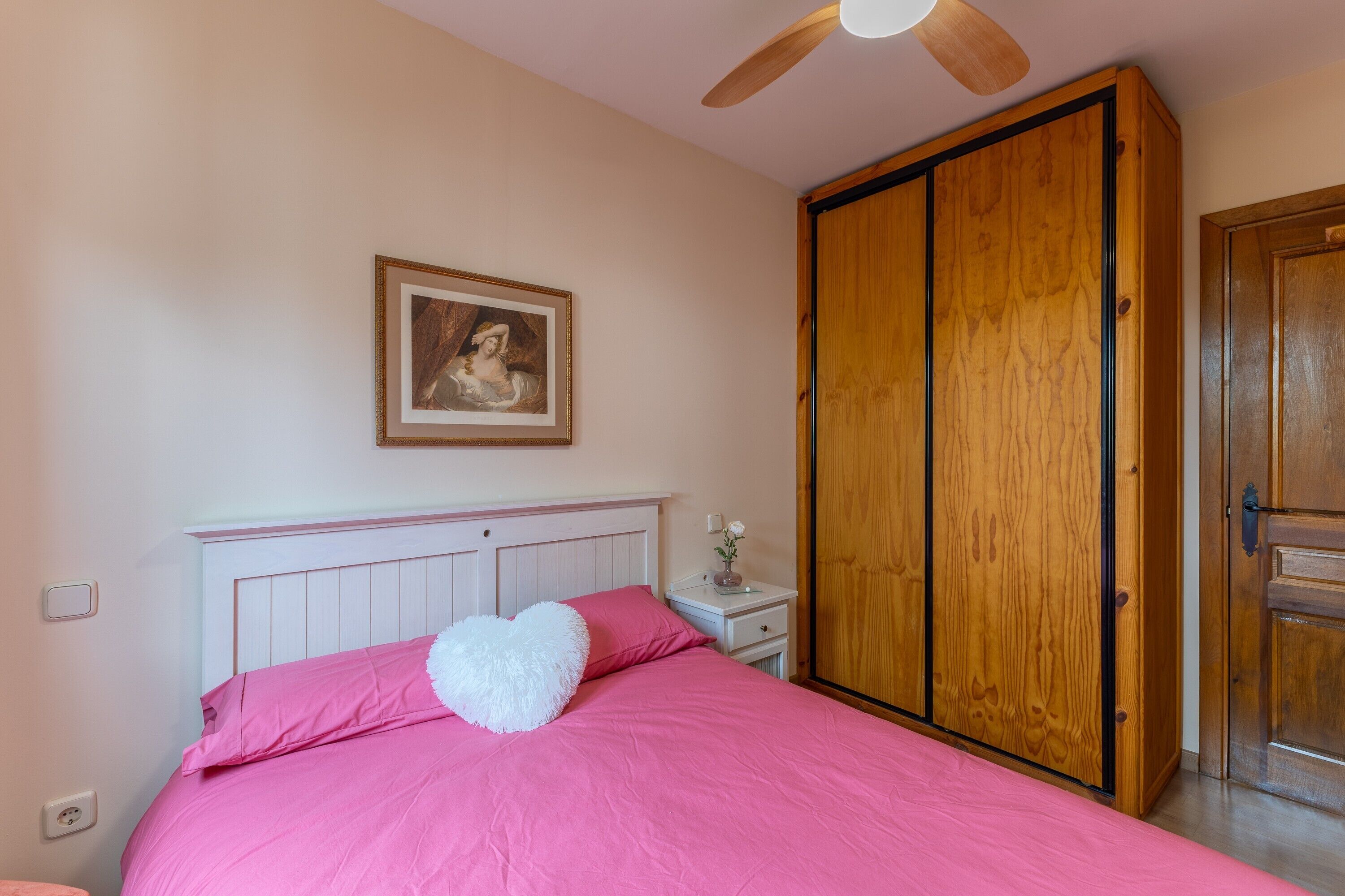 Un dormitorio con una cama, un armario de madera, una mesita de noche con un florero y un ventilador de techo.