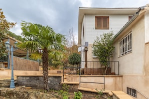 A residential building with a stone wall, a palm tree, and a staircase leading to a balcony.
