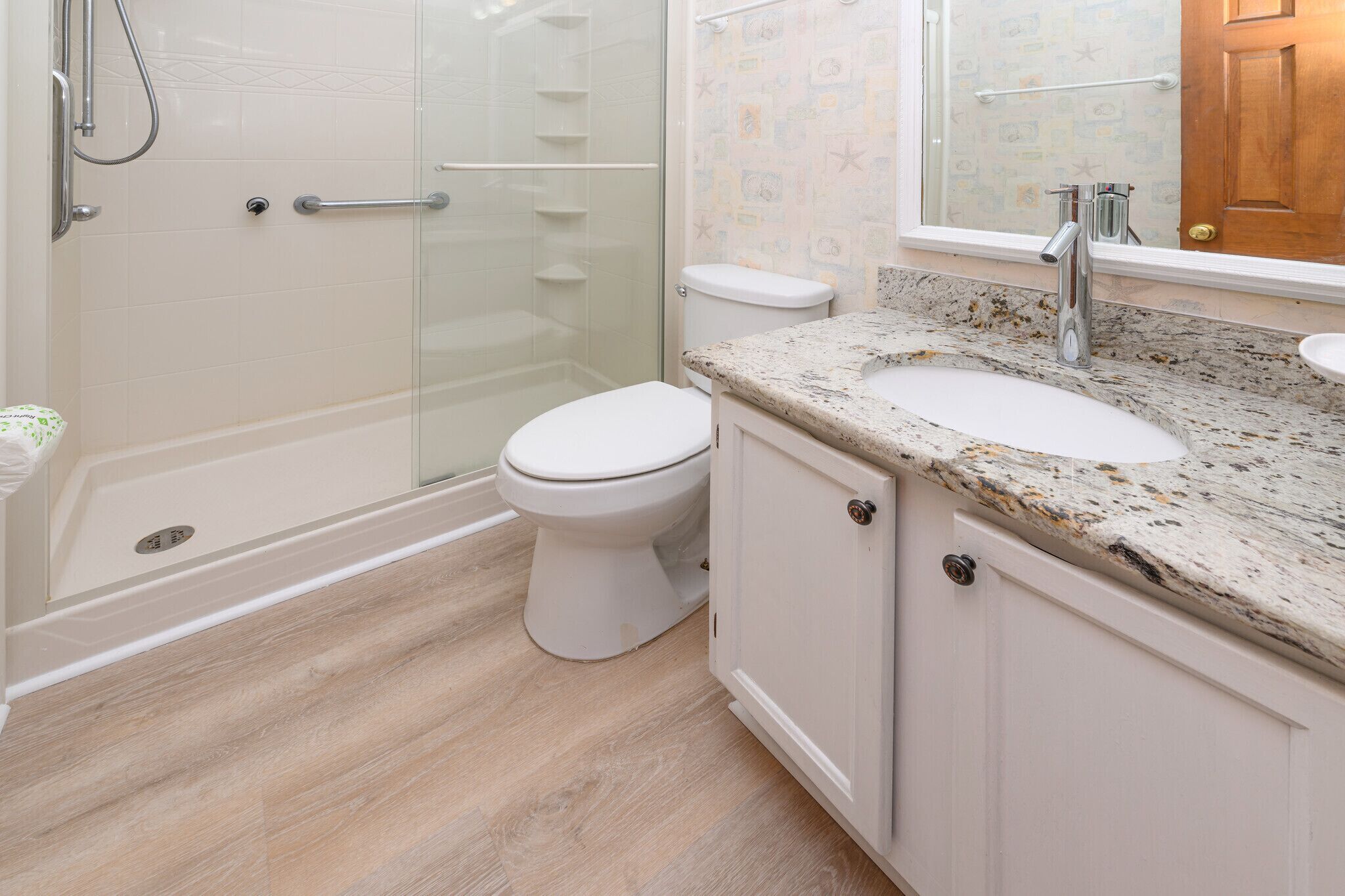 Un bany amb dutxa de vidre, vàter blanc, un lavabo amb taulell de marbre i una porta de fusta.