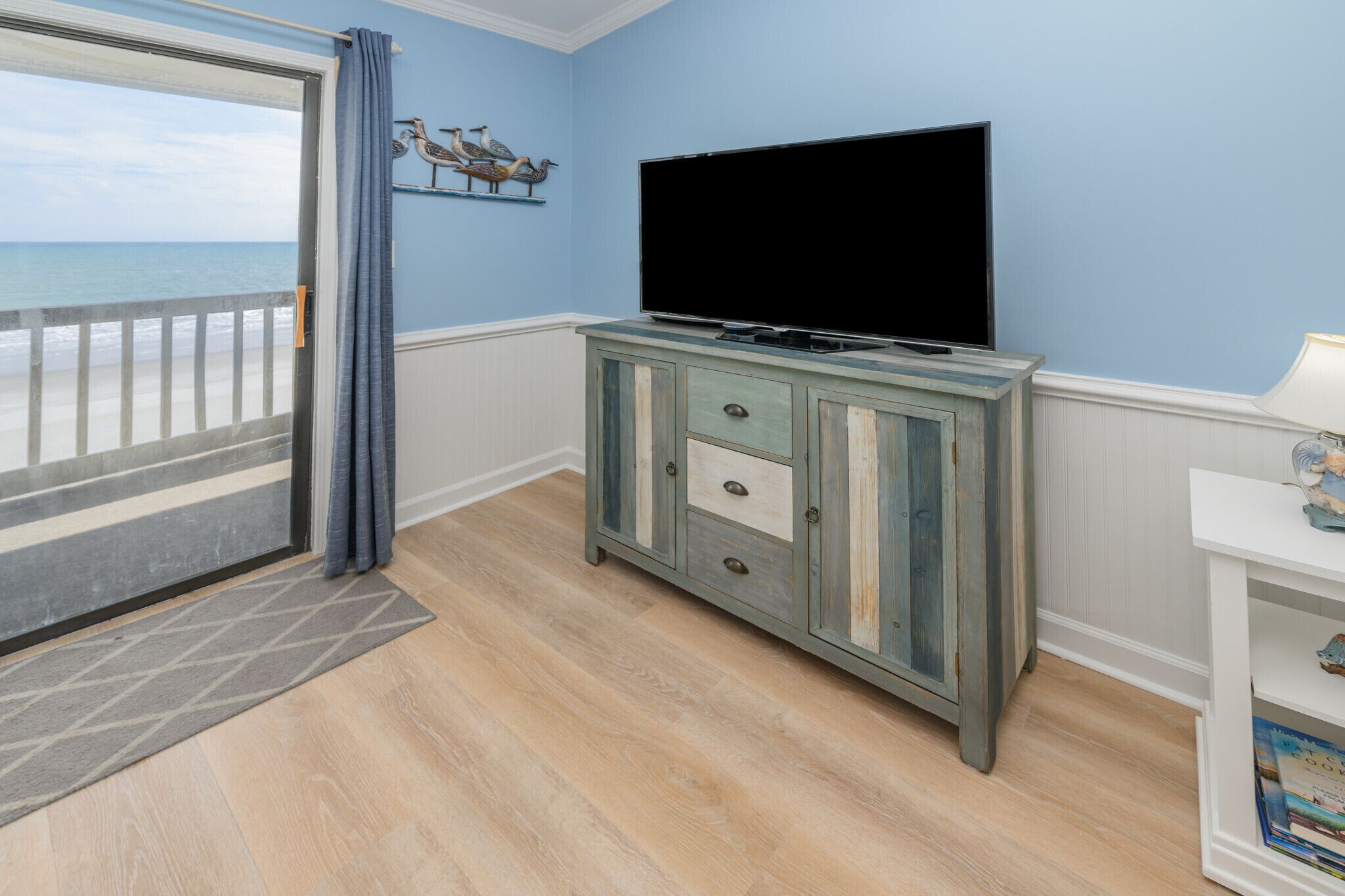 A room with a large window overlooking the beach, a wooden cabinet with drawers, a flat-screen TV, and a small white shelf with books.