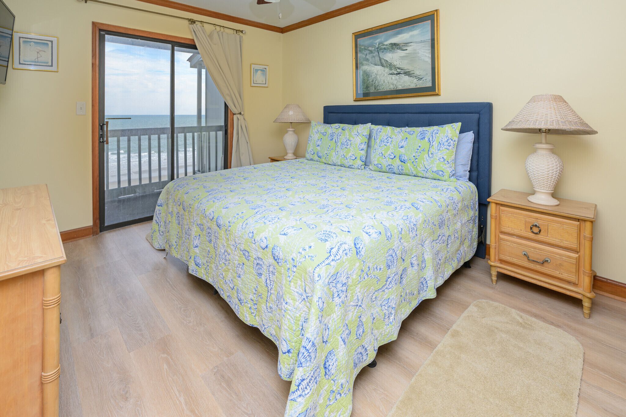 A bedroom with a bed, a nightstand, a lamp, a wooden dresser, a window with curtains, and a painting on the wall.