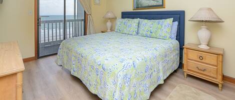 A bedroom with a bed, a nightstand, a lamp, a wooden dresser, a window with curtains, and a painting on the wall.