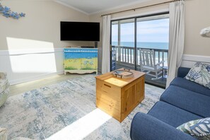 A living room with a sofa, a wooden coffee table, a TV, and a view of the ocean through a balcony. - Oceanfront, Shared Pool - Surfwatch II 404 (Surfside Beach)