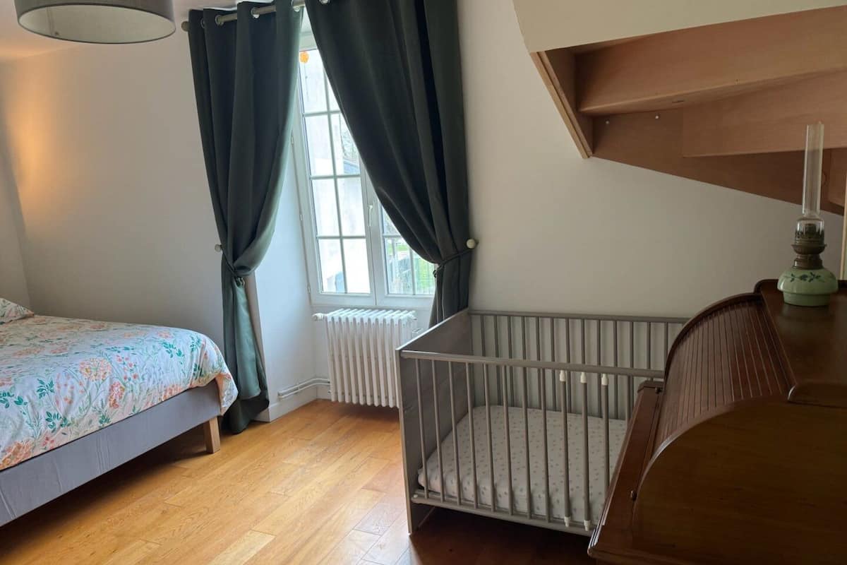 A bedroom with a wooden floor, a crib, a bed with a floral bedspread, a radiator, and a window with curtains.