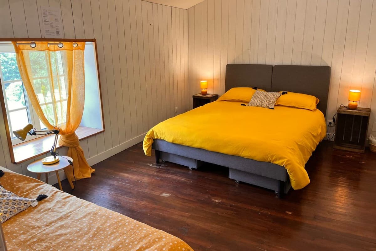 A bedroom with a bed, two bedside tables with lamps, a window with curtains, and a wooden floor.
