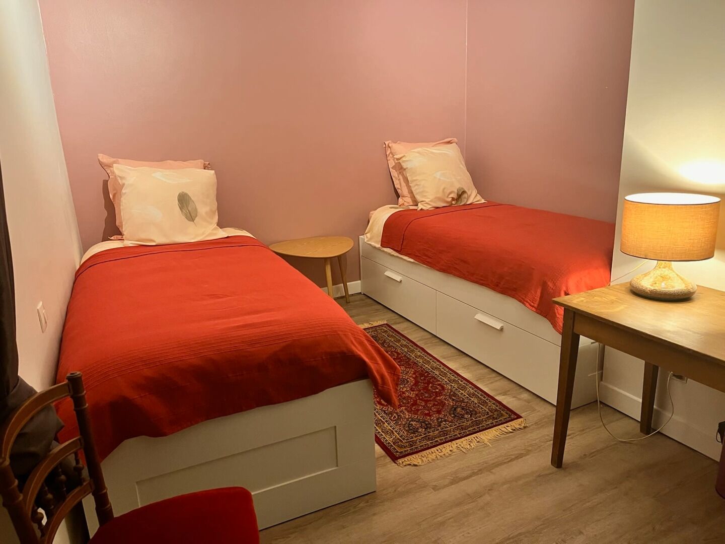 Um quarto pequeno com duas camas de solteiro, uma escrivaninha de madeira, uma cadeira e uma mesinha.