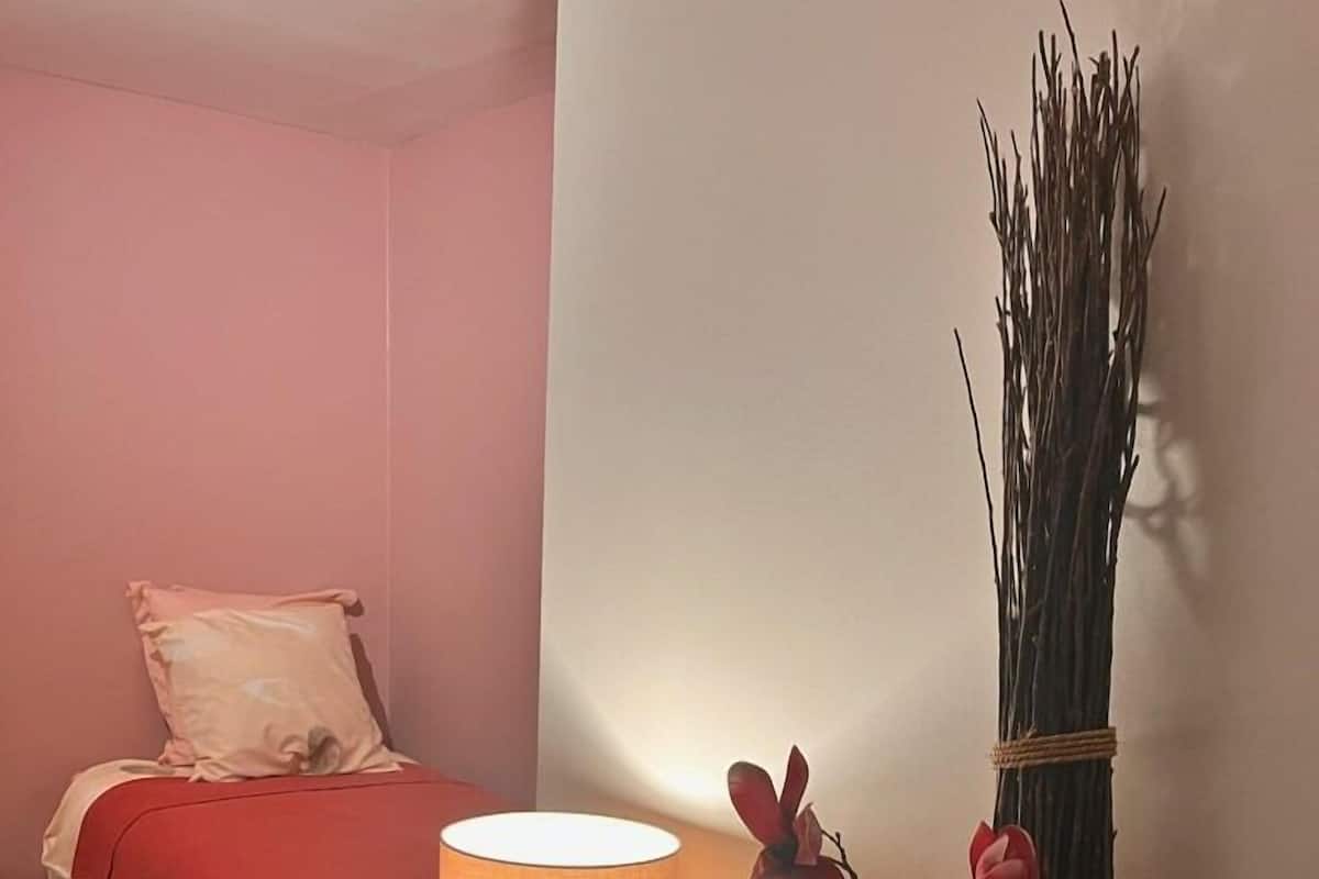 A bedroom with a bed, a bedside table with a lamp, a vase with branches, and a red rug.