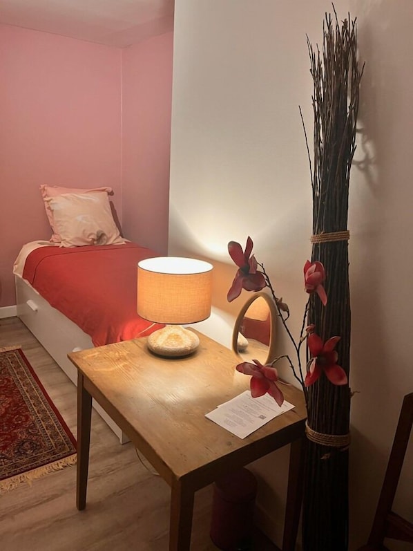 Un dormitorio con cama, mesita de noche con lámpara, un jarrón con ramas y una alfombra roja.