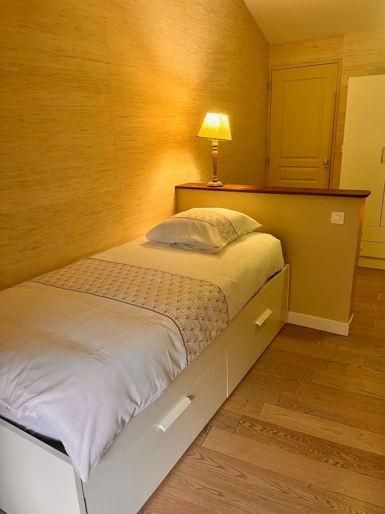 A small, neatly made bed with a patterned bedspread and pillows, a bedside table with a lamp, and a wooden floor.