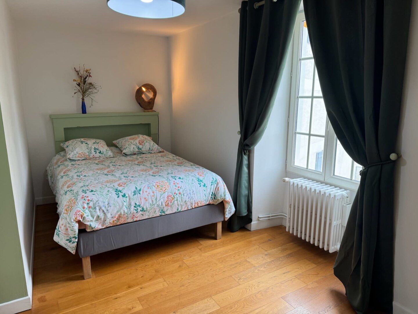 A bedroom with a bed, a radiator, a window with curtains, and a vase with dried flowers on the nightstand.