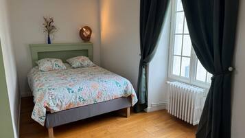 Ein Schlafzimmer mit einem Bett, einem Heizkörper, einem Fenster mit Vorhängen und einer Vase mit getrockneten Blumen auf dem Nachttisch.