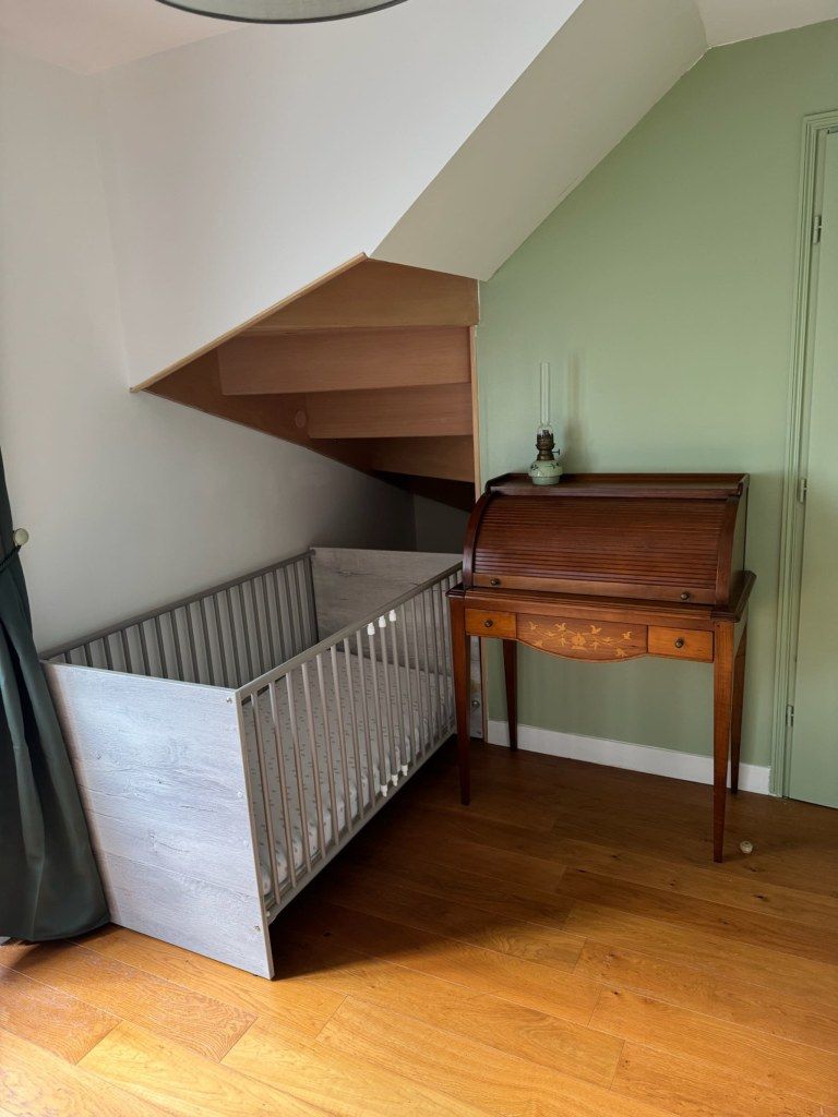 Une pièce avec un berceau en bois, une petite table avec une lampe et un plafond en pente.