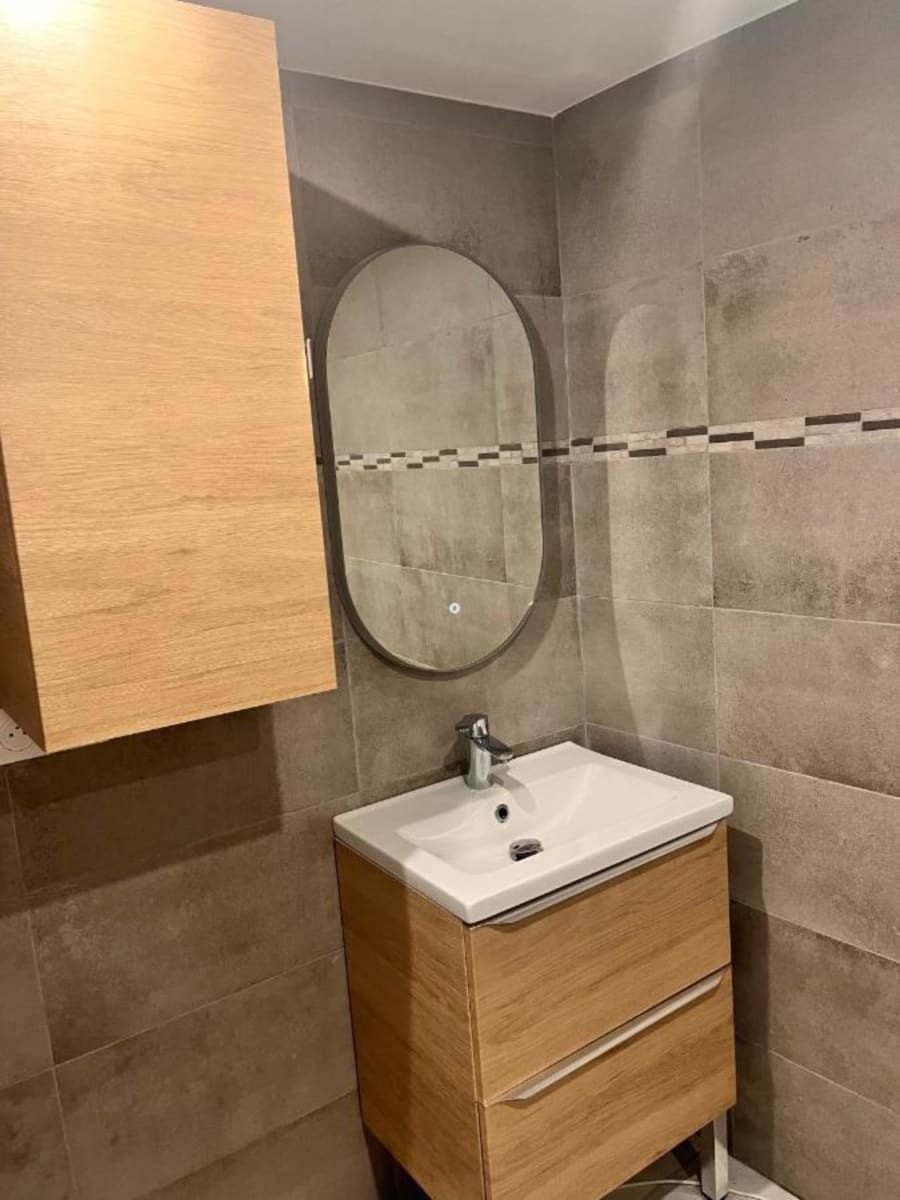 Une salle de bain avec un meuble en bois, un miroir rond et un lavabo blanc avec un robinet.