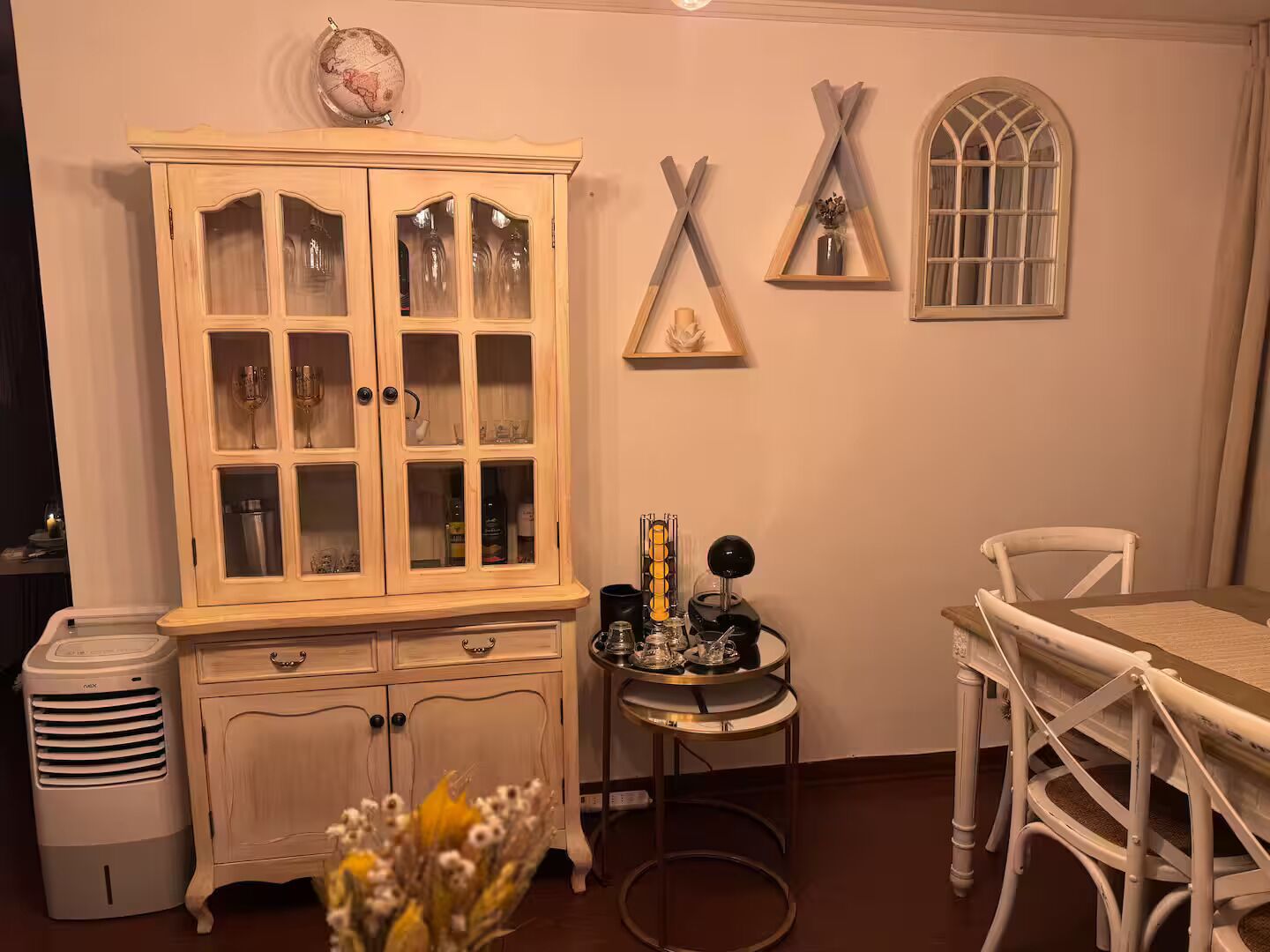 A room with a wooden cabinet, a small table with a lamp, a globe on a shelf, and a window with a decorative frame.