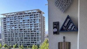 A balcony with a view of a high-rise building, a heart-shaped decoration, a bottle of beer, and a triangular sign with text. - Excelente Ubicación y Confort. (Las Condes)