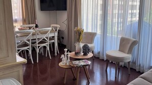 A living room with a sofa, a dining table with chairs, a TV mounted on the wall, and a Buddha statue on a side table. - Excelente Ubicación y Confort. (Las Condes)