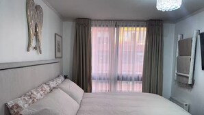 A bedroom with a large bed, a chandelier, a window with curtains, and a radiator. - Excelente Ubicación y Confort. (Las Condes)