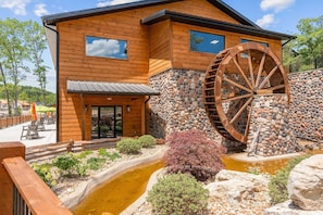 A wooden building with a large water wheel, a stone wall, and a small pond with vegetation.