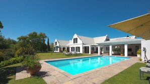 Une piscine avec un espace patio, entourée d’une pelouse bien entretenue et d’une grande maison à l’extérieur blanc, dotée d’un patio couvert.