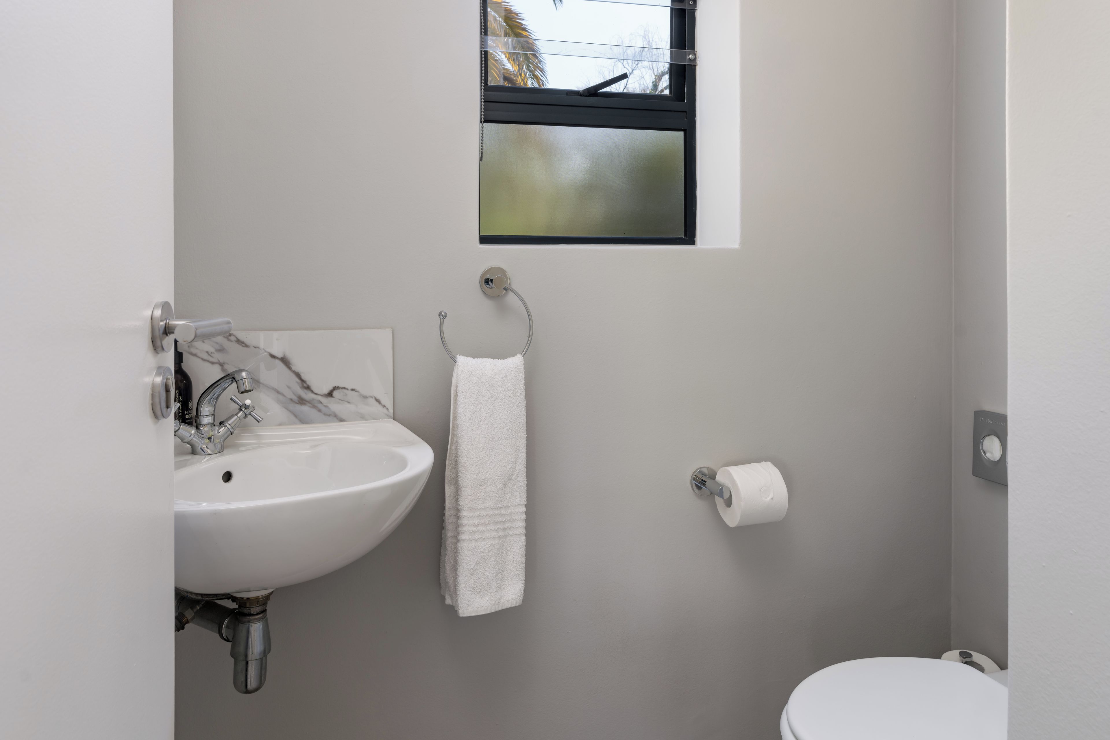 Un bagno con un lavandino bianco, un asciugamano appeso a un portasciuga metallico, un water e una piccola finestra vista alberi.